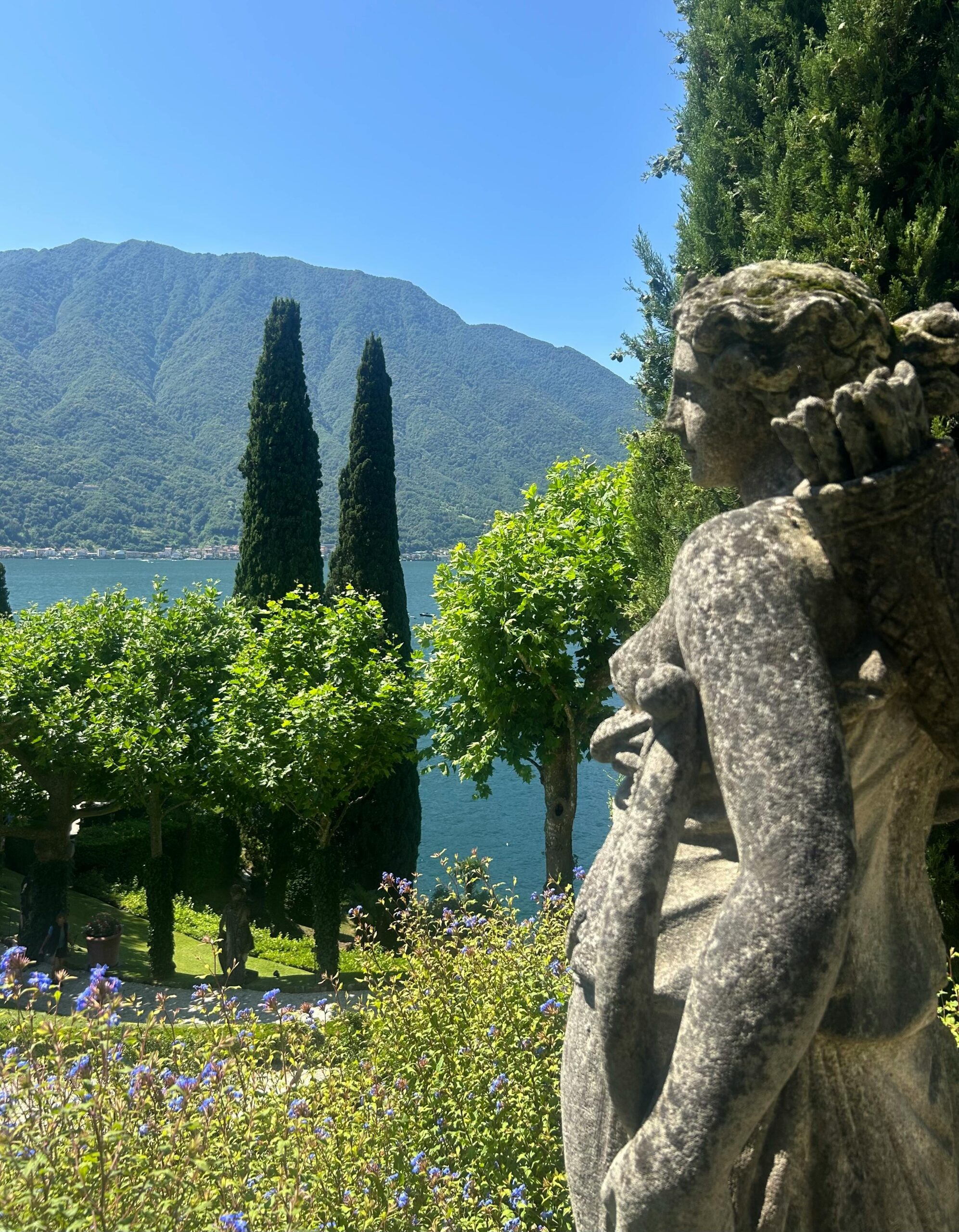 Benvenuto A peaceful lakeside landscape with a classical statue, vibrant greenery, and mountains in the background.